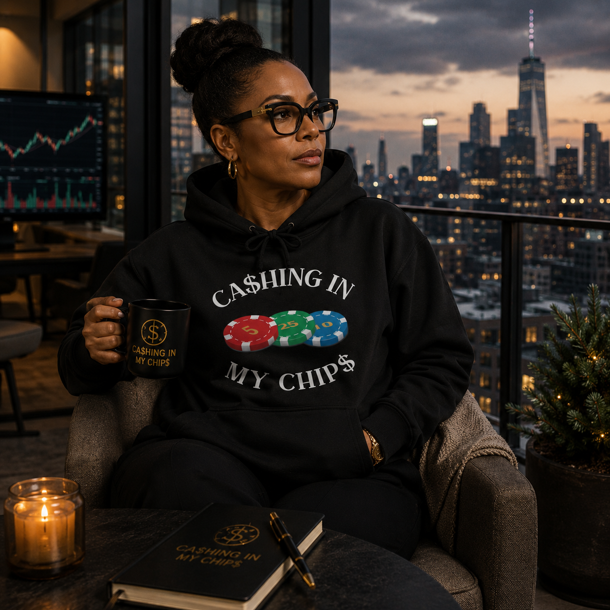 African American woman wearing a black Ca$hing In My Chip$ hoodie with colorful poker chip design, seated in a luxury high-rise overlooking the New York City skyline while holding a branded coffee mug.