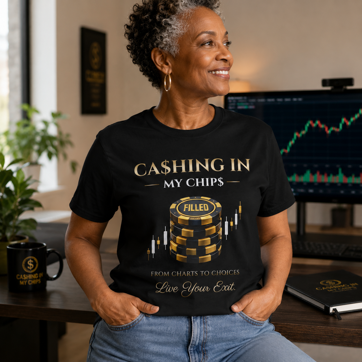 Older African American woman wearing a black Ca$hing In My Chip$ “Filled” graphic T-shirt in a modern trading office with stock charts displayed on a monitor in the background.